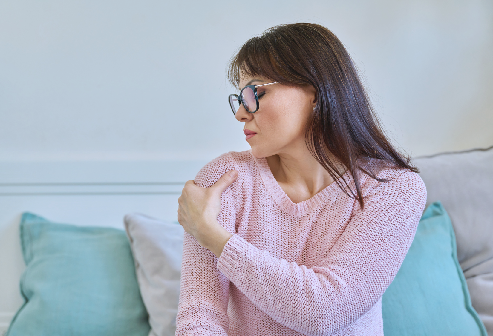 Recommended Bra for Shoulder Surgery Recovery: A woman sitting on a light blue couch, wearing a pink sweater, feeling pain in her shoulder and holding it with her left hand.