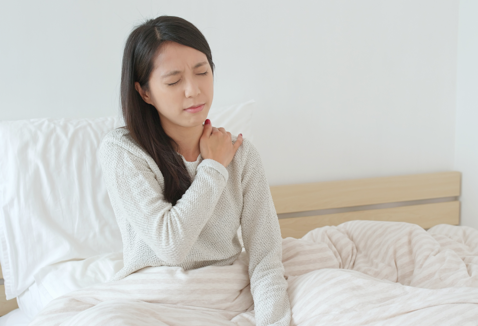 How to Put on a Bra After Shoulder Surgery: A woman with shoulder pain is holding her left shoulder with her hand while lying in bed.