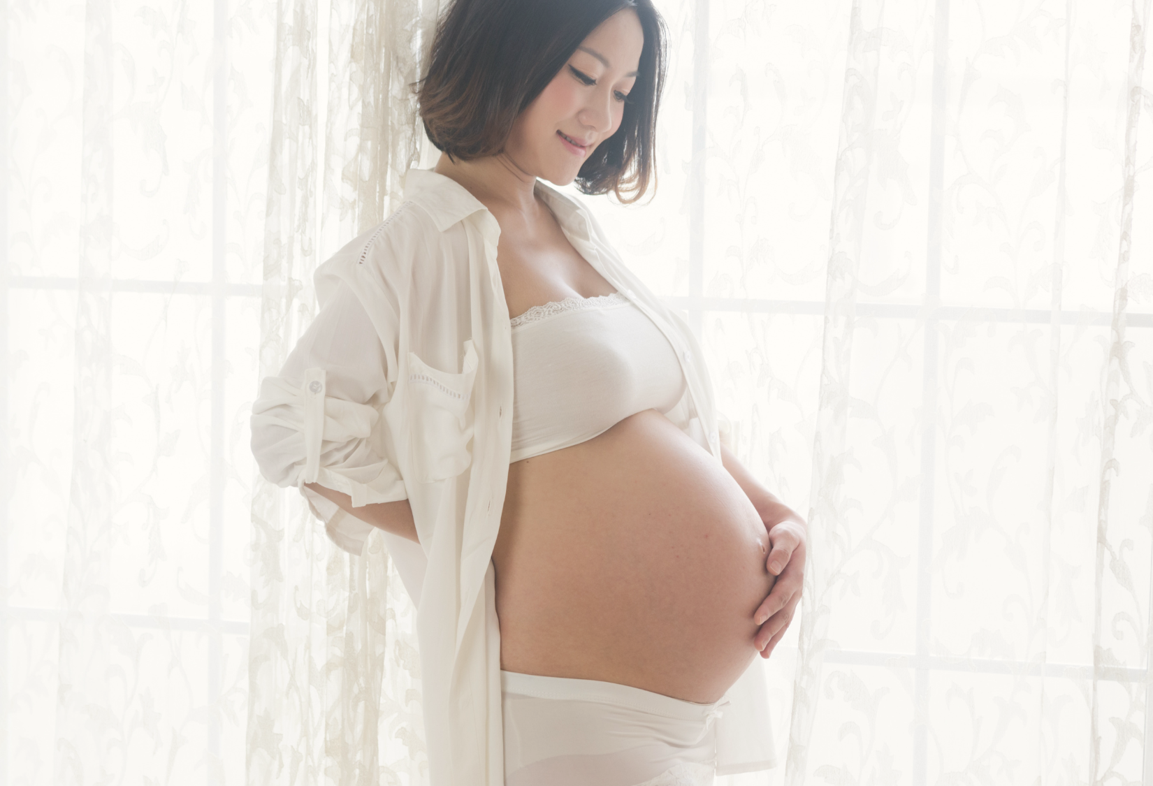 Asian woman dressed in white showing her pregnant belly and holding it while smiling