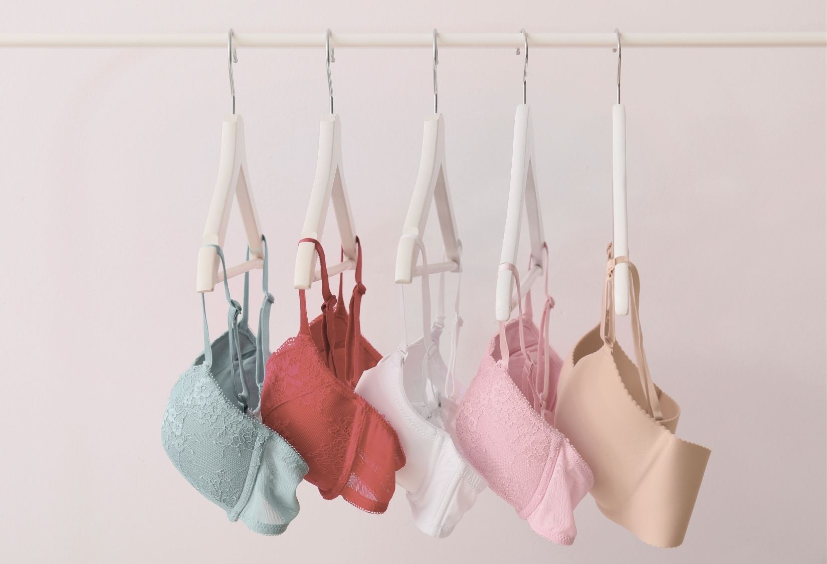 Bra fabrics: A variety of bras in pastel colors, including lace and smooth styles, hanging neatly on a clothing rack against a light pink background.