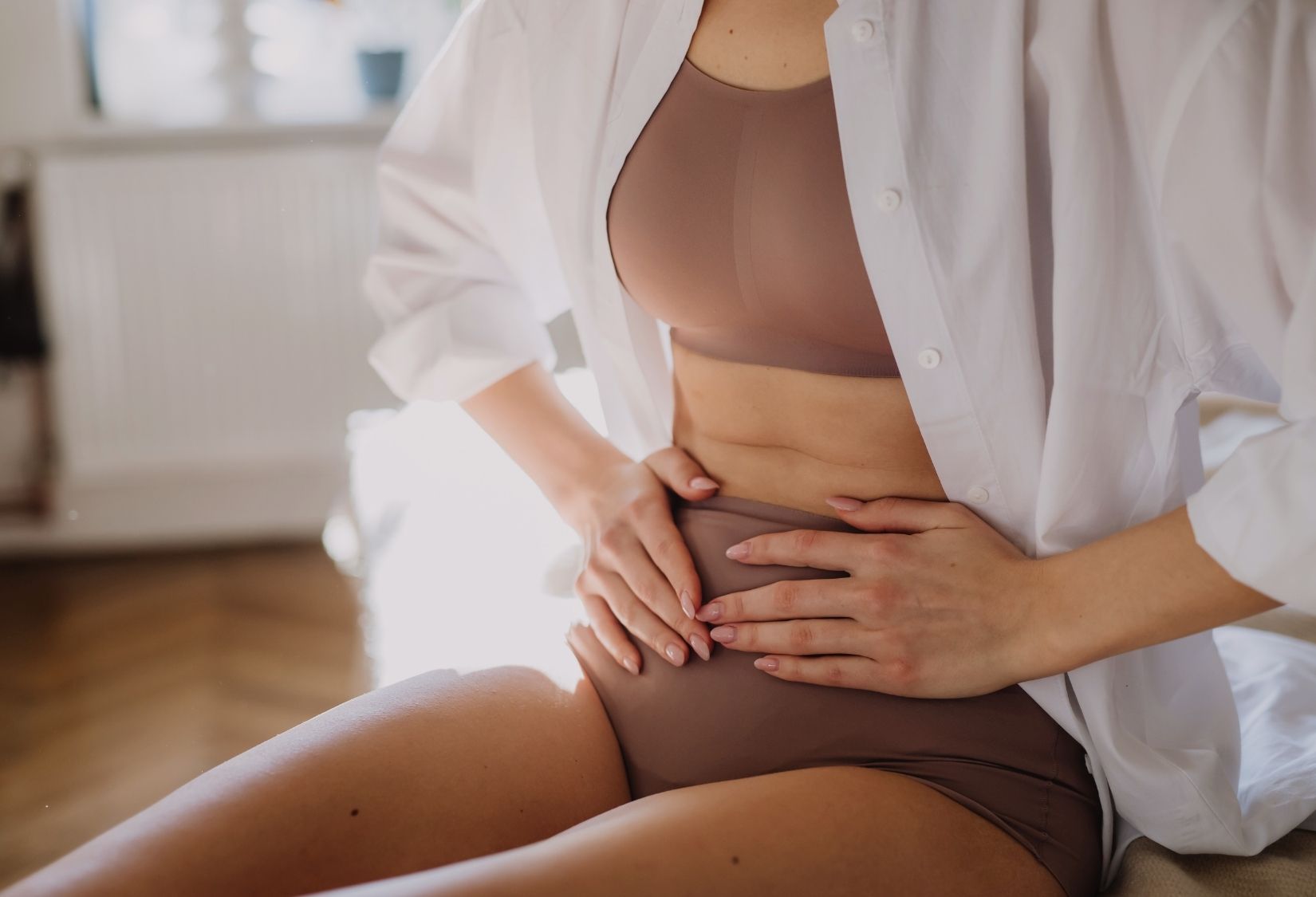 Fibromyalgia and Periods:  A woman experiencing fibromyalgia and period discomfort, sitting with hands on lower abdomen in a neutral-colored bra and underwear.