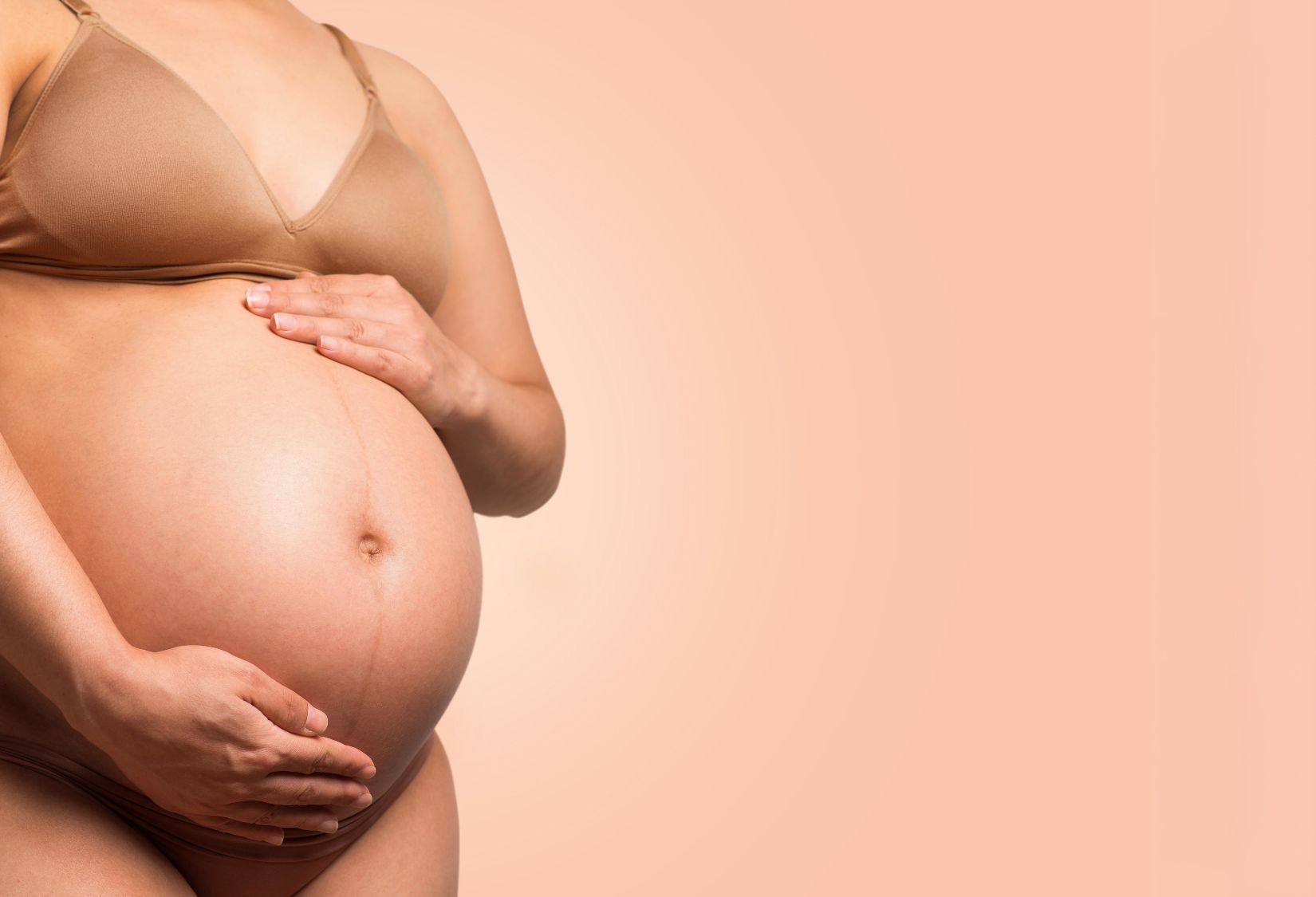 Lupus and Pregnancy: A pregnant woman wearing a beige bra holds her bare belly with both hands against a soft peach background.