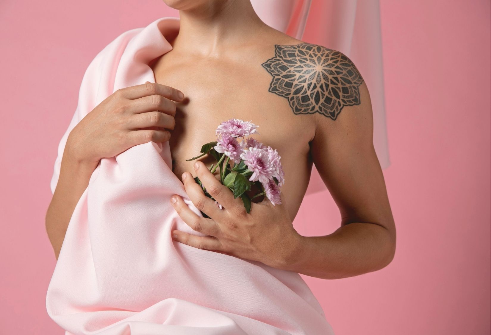 Bras Post Mastectomy: Person gently holding flowers close to their chest, symbolizing healing and renewal after a mastectomy, with a soft pink fabric draped across their body and a floral tattoo on the shoulder, set against a pink background.