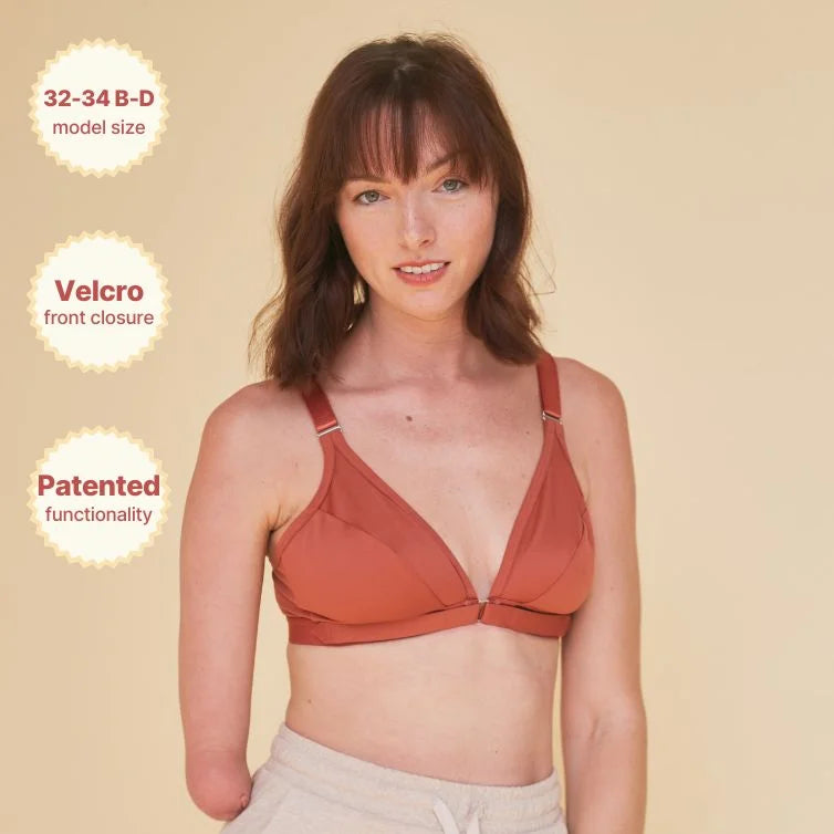 Young woman wearing the Springrose one handed bra in terracotta while lightly smiling. The image highlights the size, the velcro front closure, and patented functionality.